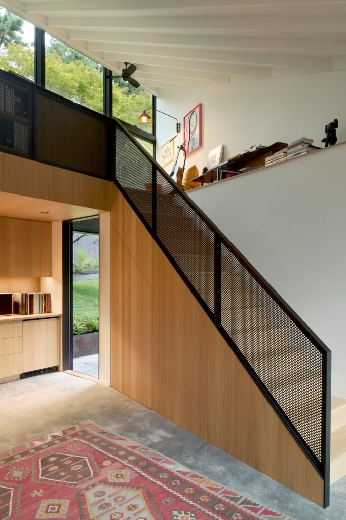 Wooden interior staircase with black perforated metal mesh railing leading to an upper mezzanine in a minimalist home office.