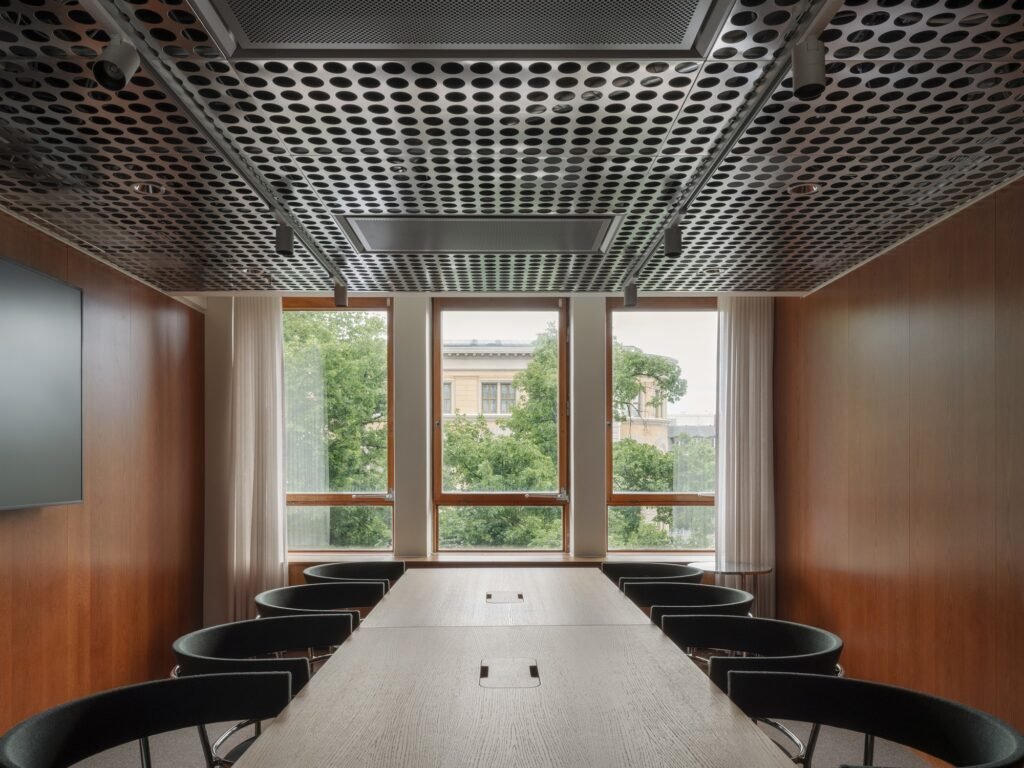 Executive boardroom table in Folkets Hus with black armchairs, warm wood wall panelling, industrial metal ceiling grid, and view to green trees through tall windows.