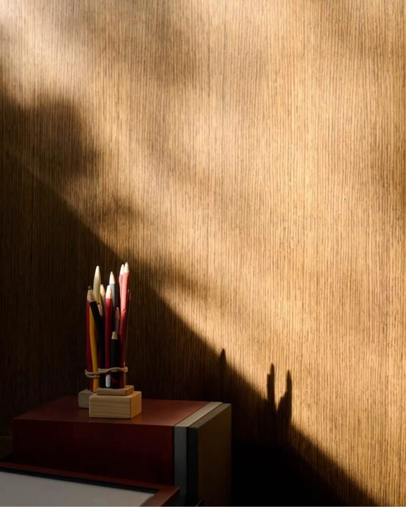Interior detail shot of natural light and shadow patterns casting across vertical grain wood wall cladding with art supplies on a ledge.