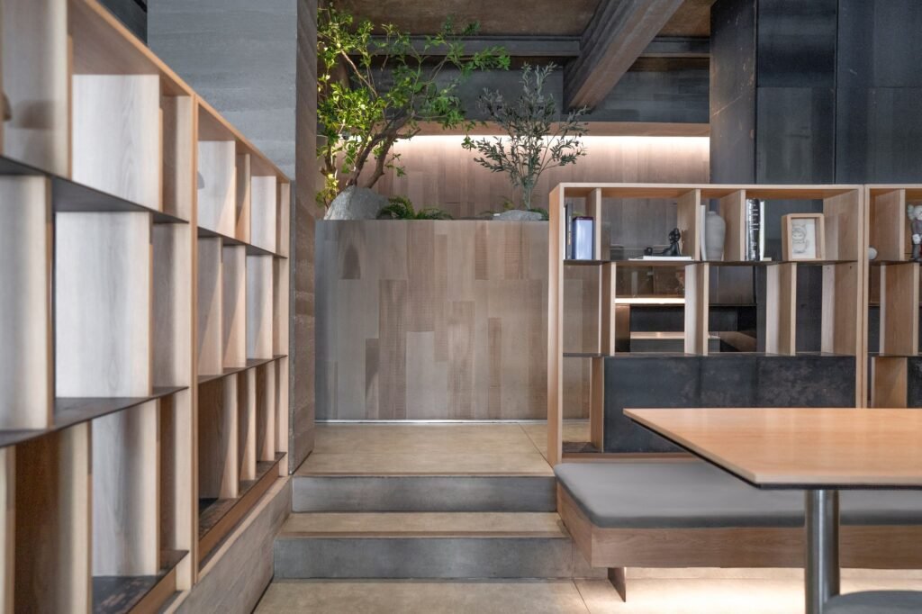 Cafe interior seating area with light wood shelving units, indoor plants, and concrete stairs.