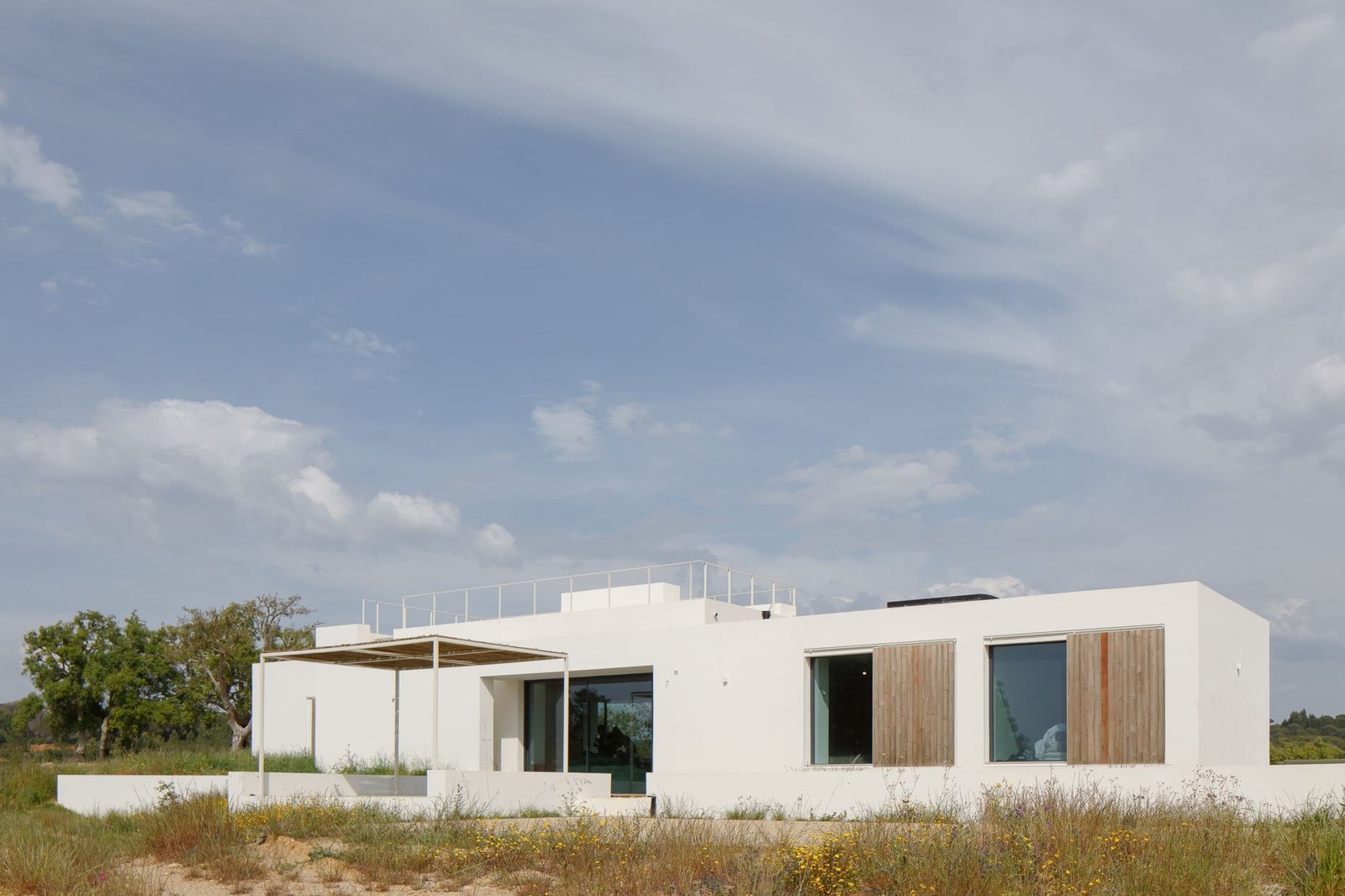 Modern white single-story residential volume of Melides MG House featuring wooden window shutters and natural terrain vegetation.