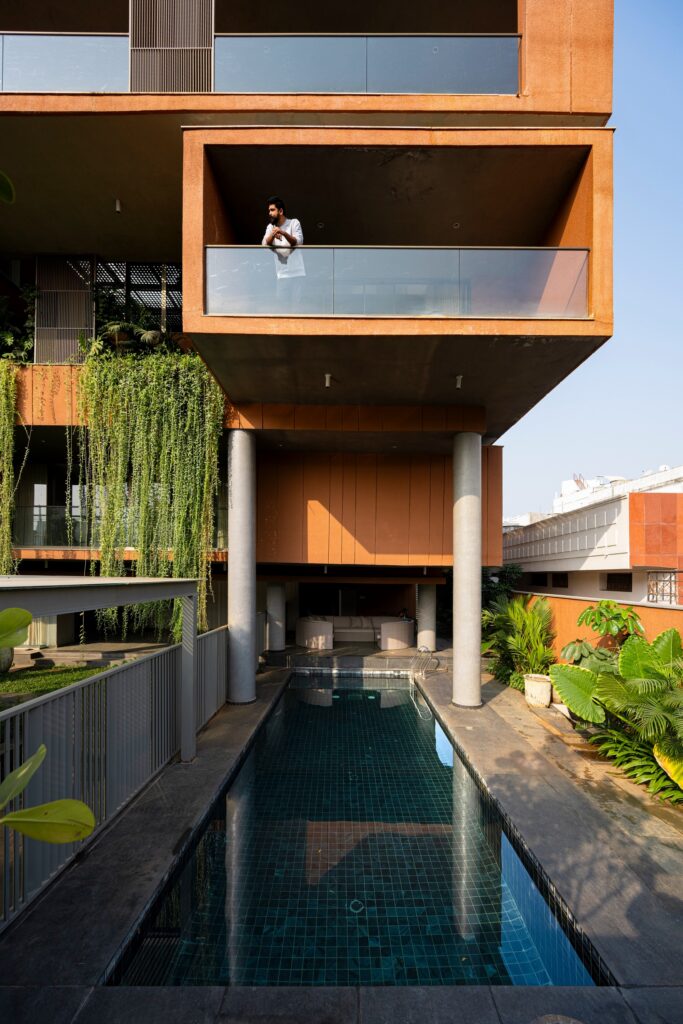 Ground floor lap pool set underneath cantilevered upper balconies supported by concrete columns with a man standing on a balcony above.