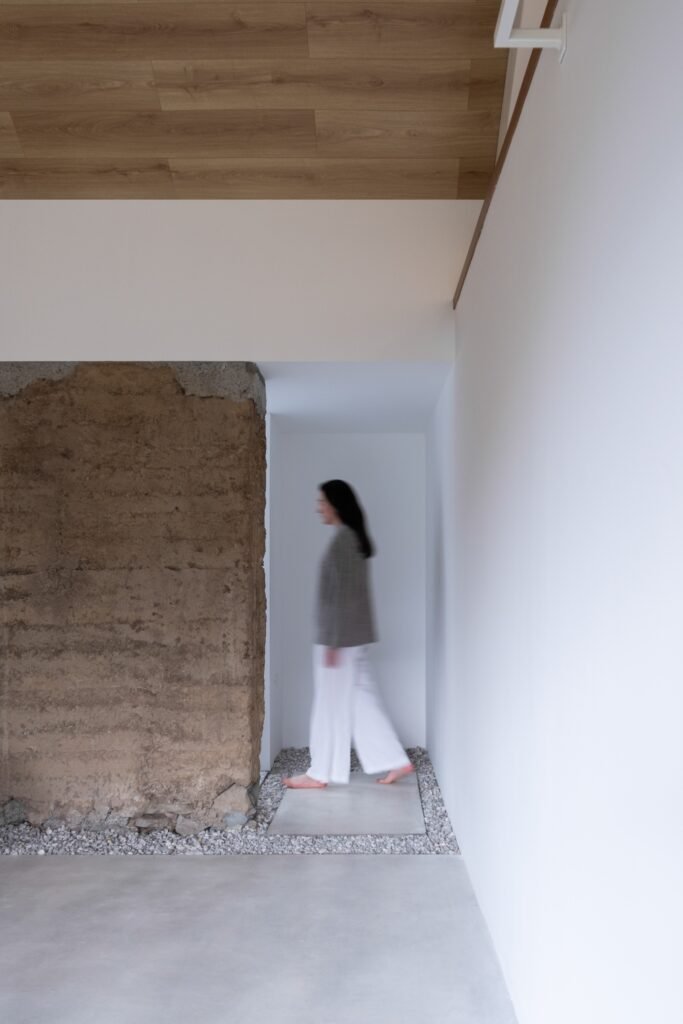 A person walking through a bright hallway lined with white walls, a light concrete floor, gravel border, and a raw rammed earth wall segment.