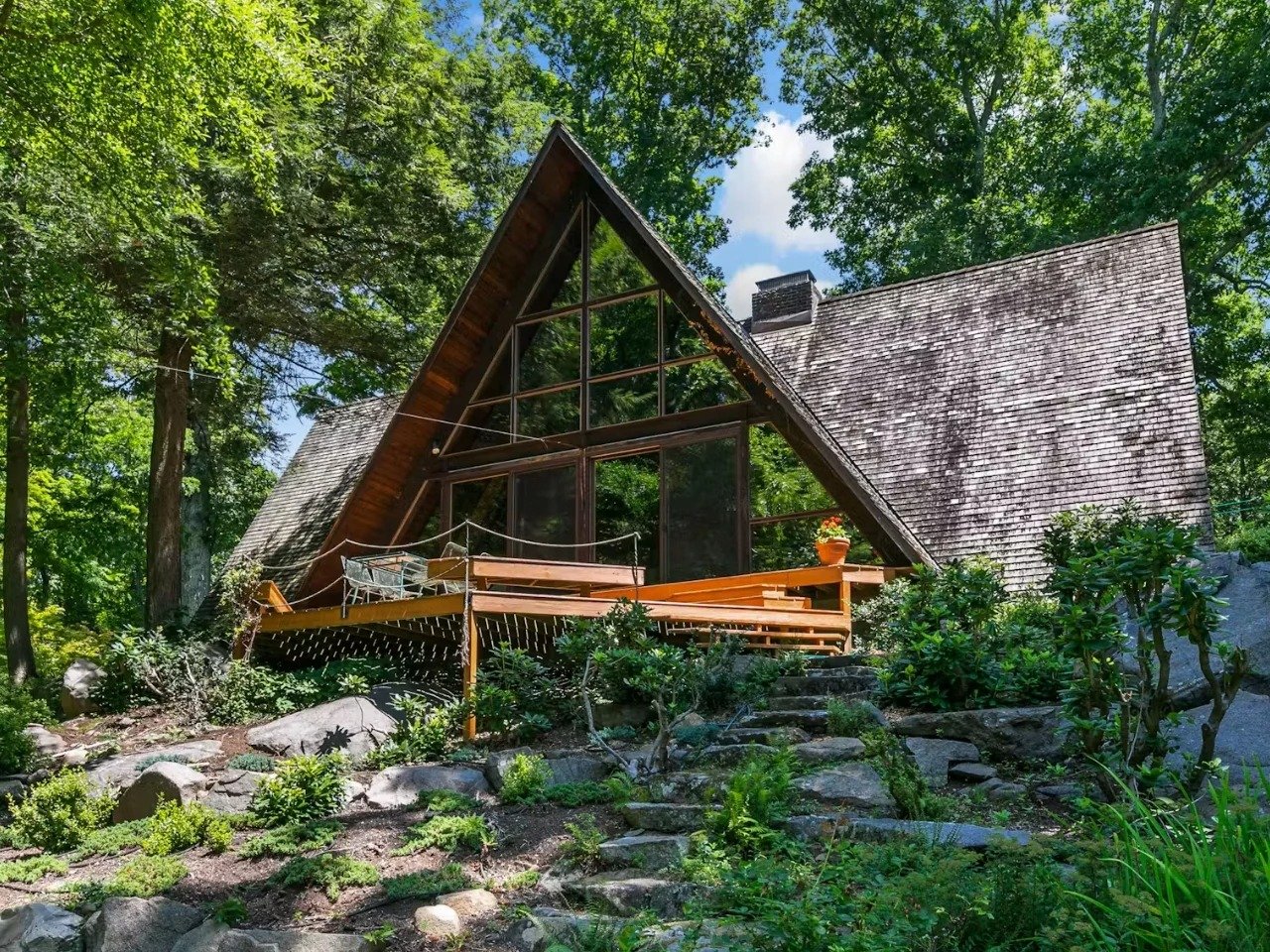 A striking exterior view of a large A-frame house in Yorktown Heights, featuring a steep shingled roof, extensive floor-to-ceiling glass windows on multiple levels, and a large wooden deck. The house is nestled into a dense, green forest under a blue sky, with a stone retaining wall and landscaped plantings in the foreground.