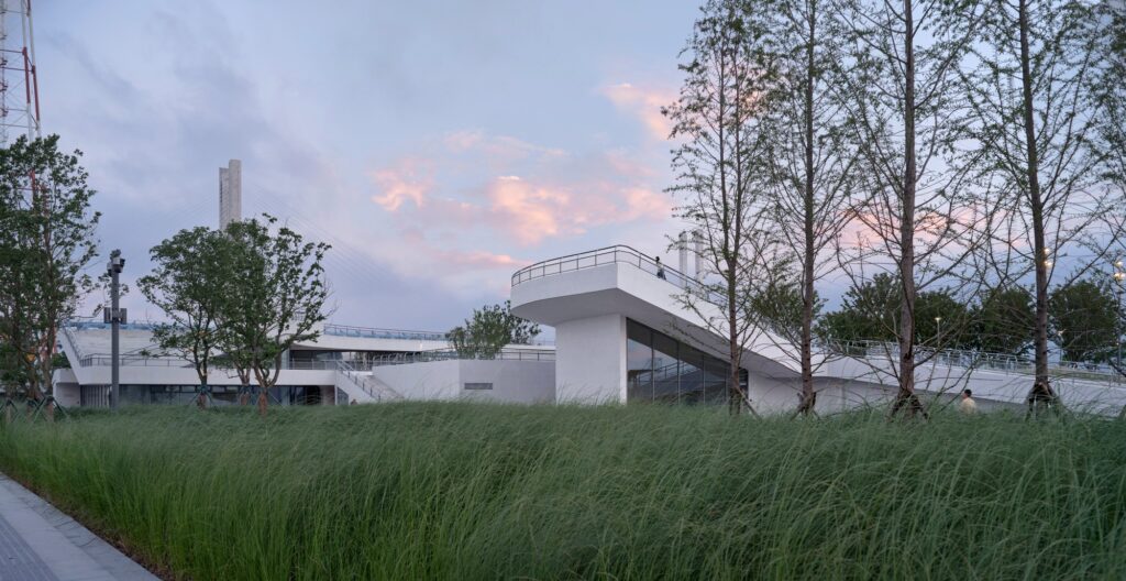 White architectural viewing platform extending over wild green grasses under a cloudy evening sky, flanked by thin trees.