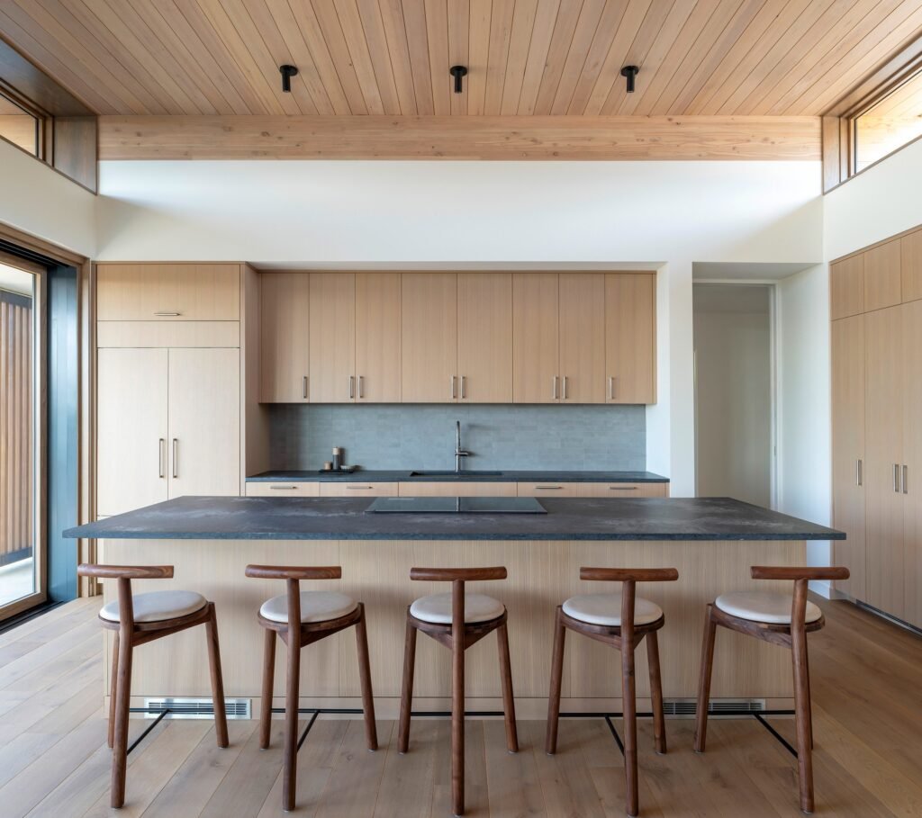 Frontal view of a modern kitchen island with five wooden barstools, wood cabinetry, and a slatted timber ceiling in Veil House.