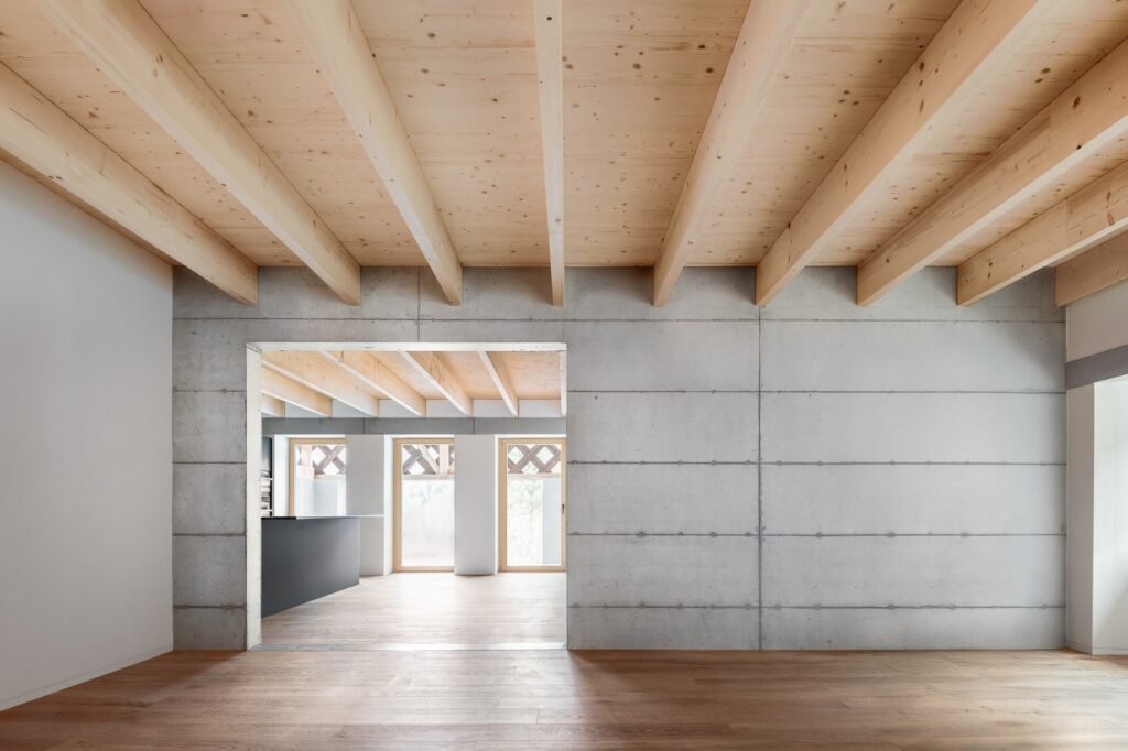Spacious interior room with exposed fair-faced concrete wall, wide doorway opening into a sunlit living space, oak flooring, and exposed timber ceiling joists.
