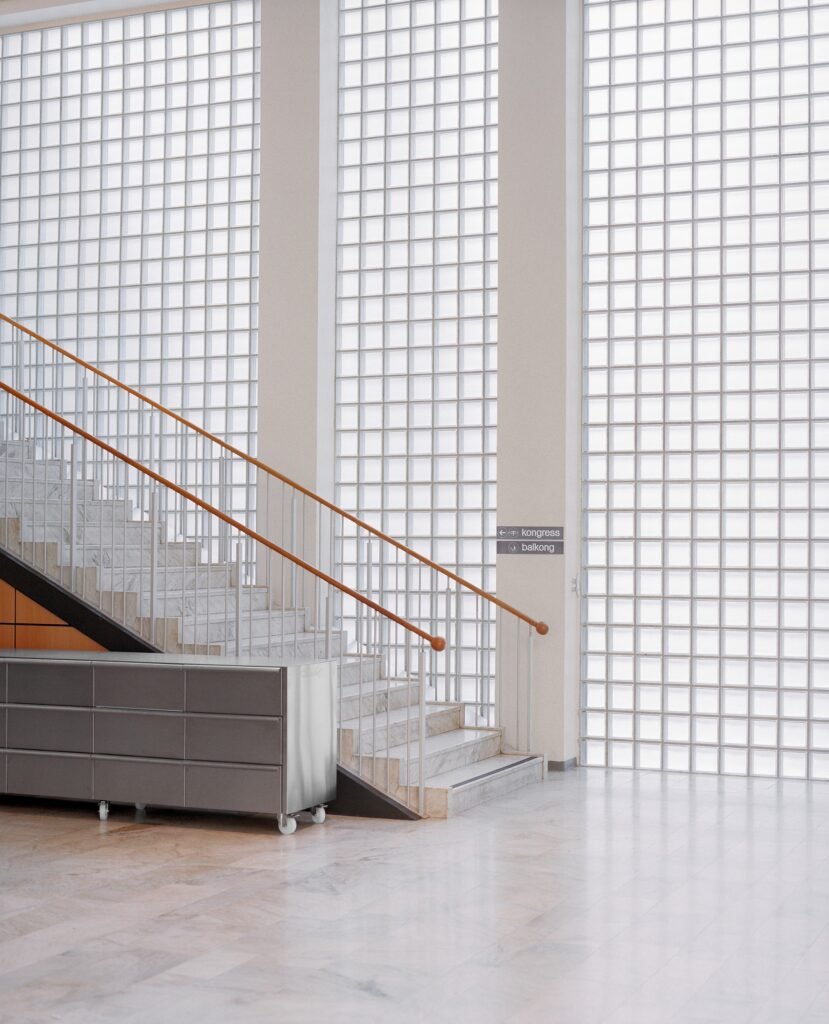 Preserved marble staircase with wooden handrail against a massive glass block facade, featuring a stainless steel storage module on wheels at Folkets Hus.