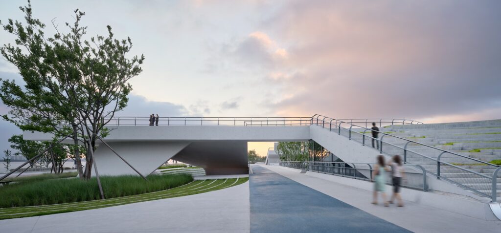 Visitors strolling along white concrete pedestrian ramps and bridges connected to broad outdoor seating steps under a soft evening sky.