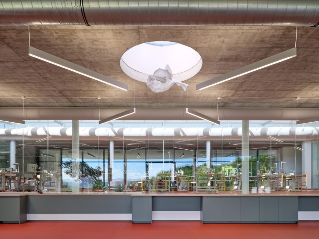 Glazed internal partition inside VAVI factory revealing sewing workstations behind glass, topped with concrete ceiling and round skylight opening.