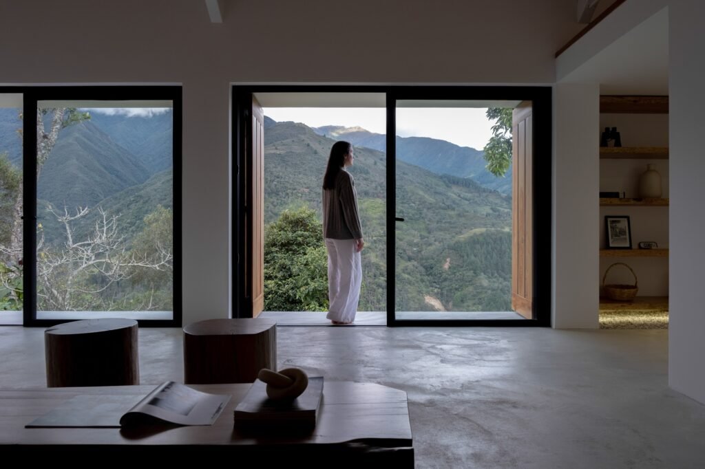 View from inside the living room toward large full-height glass doors framing a woman standing looking out at green mountain slopes.