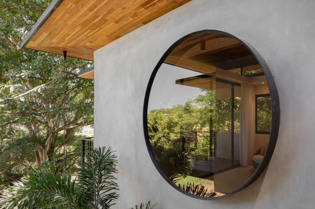 Large circular window framed in black metal set into a raw concrete exterior wall under a wooden roof overhang.