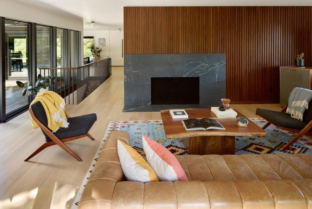 Contemporary living room with a dark stone fireplace, slatted wood feature wall, mid-century lounge chairs, and light wood flooring.