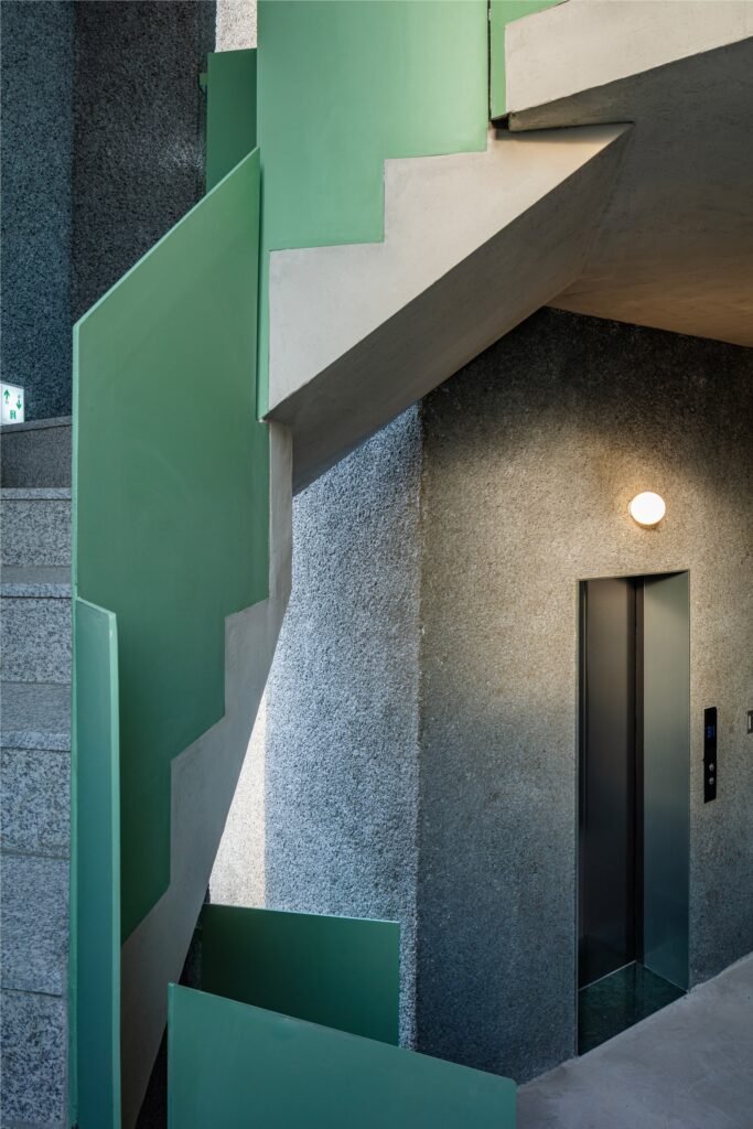 Elevator lobby and staircase detail showing green railings, rough concrete walls, and warm lighting.