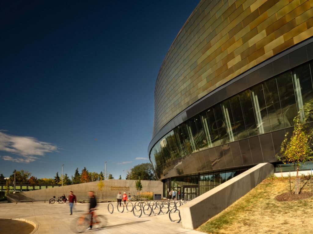 Street-level view of the main entrance to Coronation Park Sports Centre featuring bicycle racks, concrete retaining walls, and dynamic building facade.