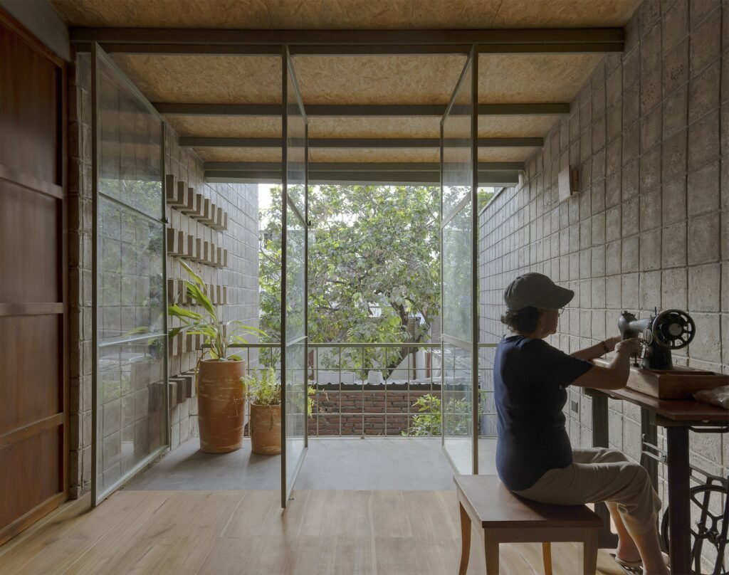 Interior space in La Colmena, where a resident works at a vintage sewing machine by large, open pivot glass windows overlooking the neighborhood trees, with natural wood flooring and concrete block walls.