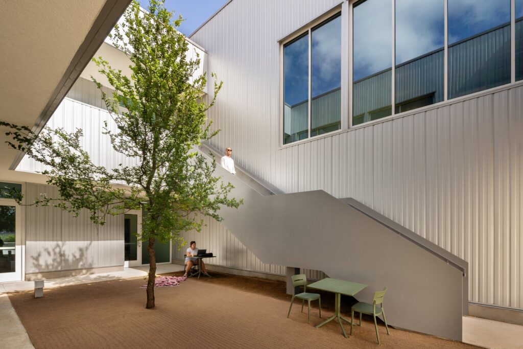 Sunlit open-air internal courtyard with a central tree, exterior steel staircase, and outdoor seating.