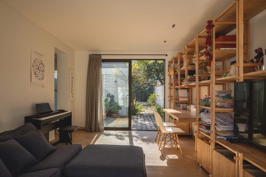 Multipurpose children's study room with wooden shelving units, piano, and glass doors leading to the garden.