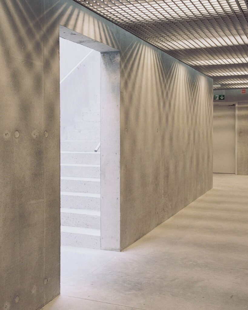 Modern interior corridor featuring raw fair-faced concrete walls, a doorway leading to stairs, and a metal grid suspended ceiling hiding mechanical services.