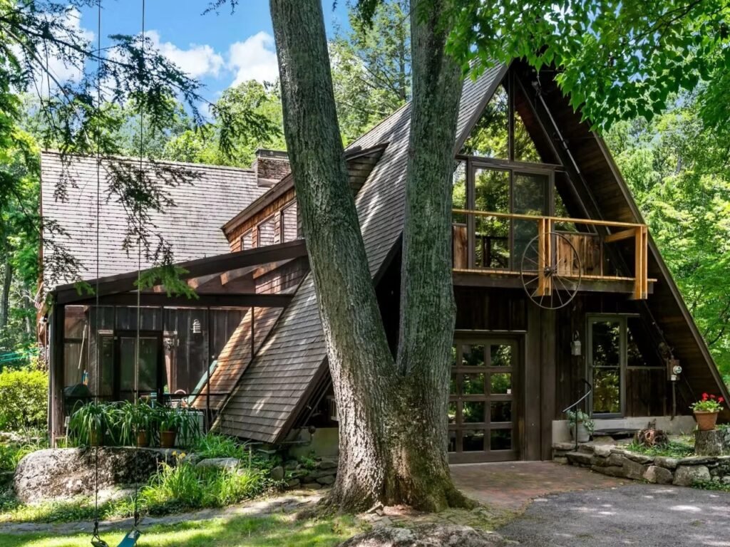 An exterior view focusing on the entrance of the A-frame house, with a large tree trunk in the foreground. A wooden balcony with a wagon wheel railing element is visible on the upper level. A grid of glass panels makes up the front door area. Green foliage is abundant.
