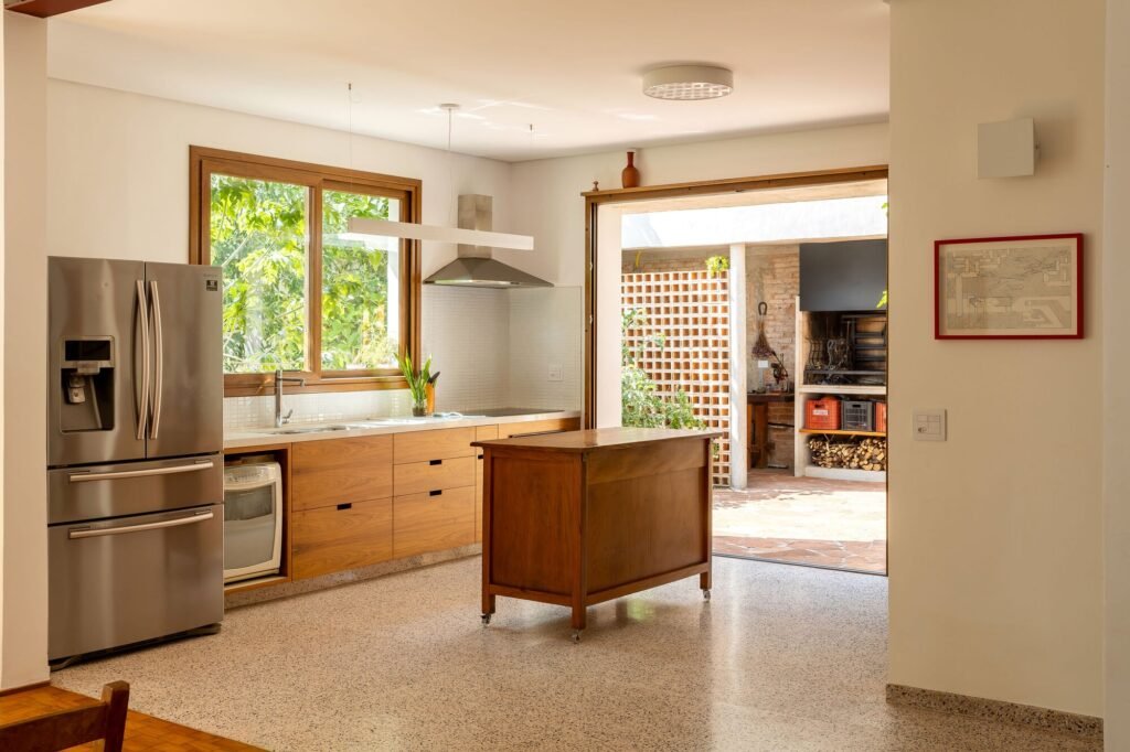 Modern kitchen with natural wood cabinetry, stainless steel appliances, polished terrazzo flooring, and glass doors opening to the courtyard.