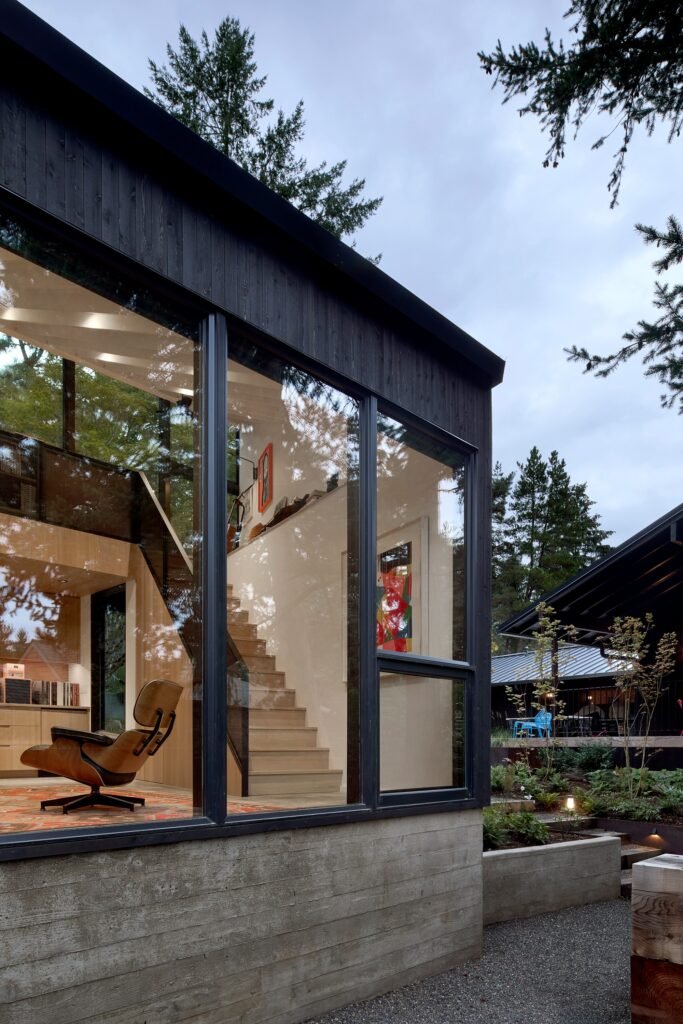 Dusk view of the detached studio exterior featuring board-formed concrete base, dark wood siding, and large glass windows.