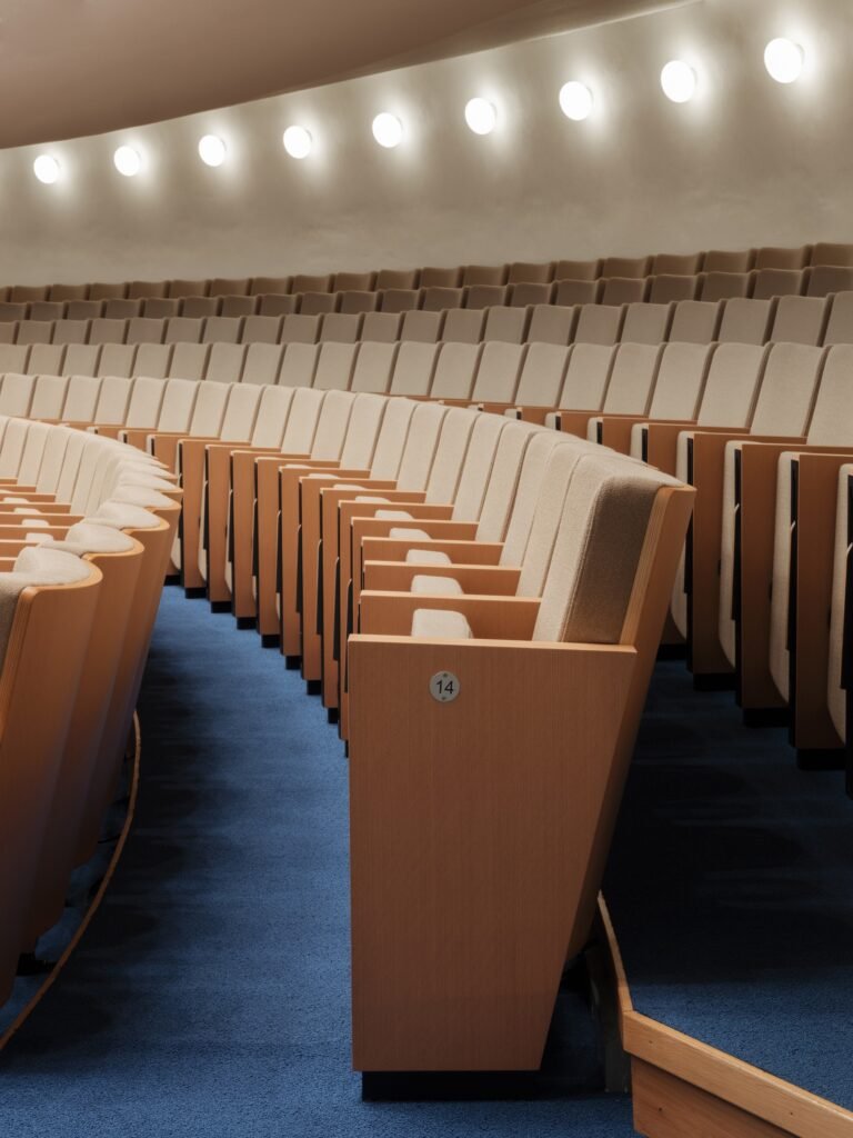 Curved rows of light-colored upholstered auditorium seating on blue carpet under soft wall sconces inside Folkets Hus auditorium.