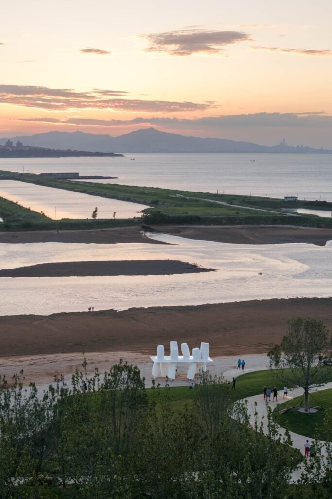 Distant elevated landscape shot showing the Coral pavilion on a sandy beach overlooking calm ocean waters and distant mountain silhouettes at sunset.