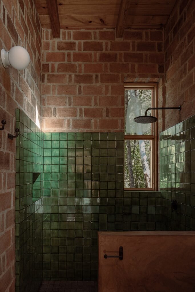 Bathroom interior with green glazed wall tiles, exposed red brickwork, a wooden window frame overlooking greenery, and a rainfall shower.