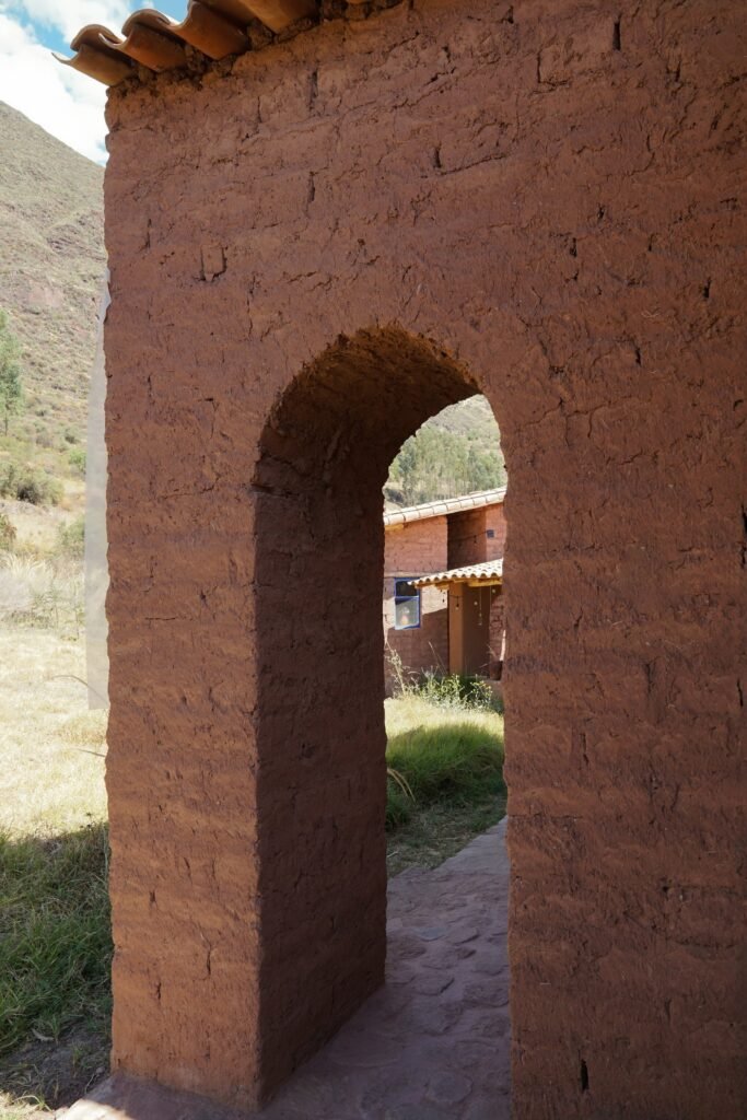 Close-up of a raw thick adobe archway leading to an outdoor walkway with adjacent buildings visible through the opening.