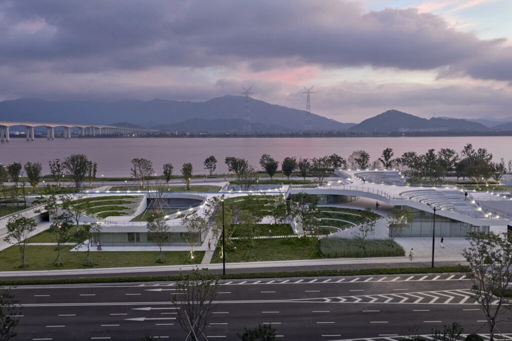 Elevated dusk shot looking across a multi-lane city road toward the illuminated green roof structures and sunken plazas of the park next to the river.