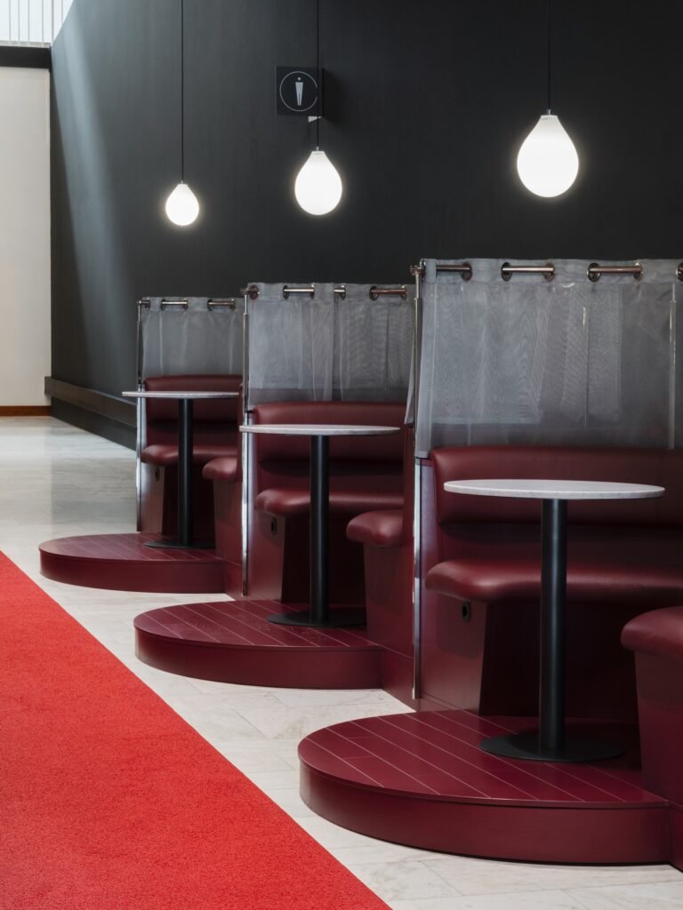 Semi-private red leather seating alcoves separated by sheer mesh curtains along a corridor with globe pendant lights in Folkets Hus.