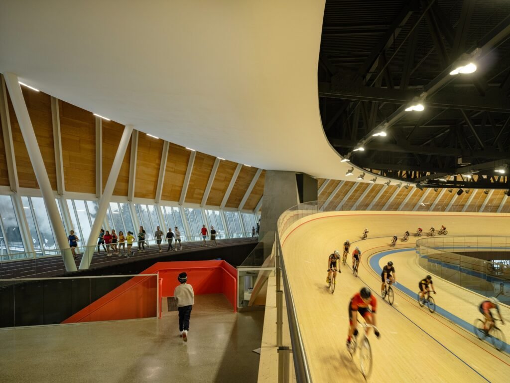 Indoor shot showing cyclists riding on the elevated wooden velodrome track alongside an inner pedestrian running track lined with timber Mass Plywood Panels.