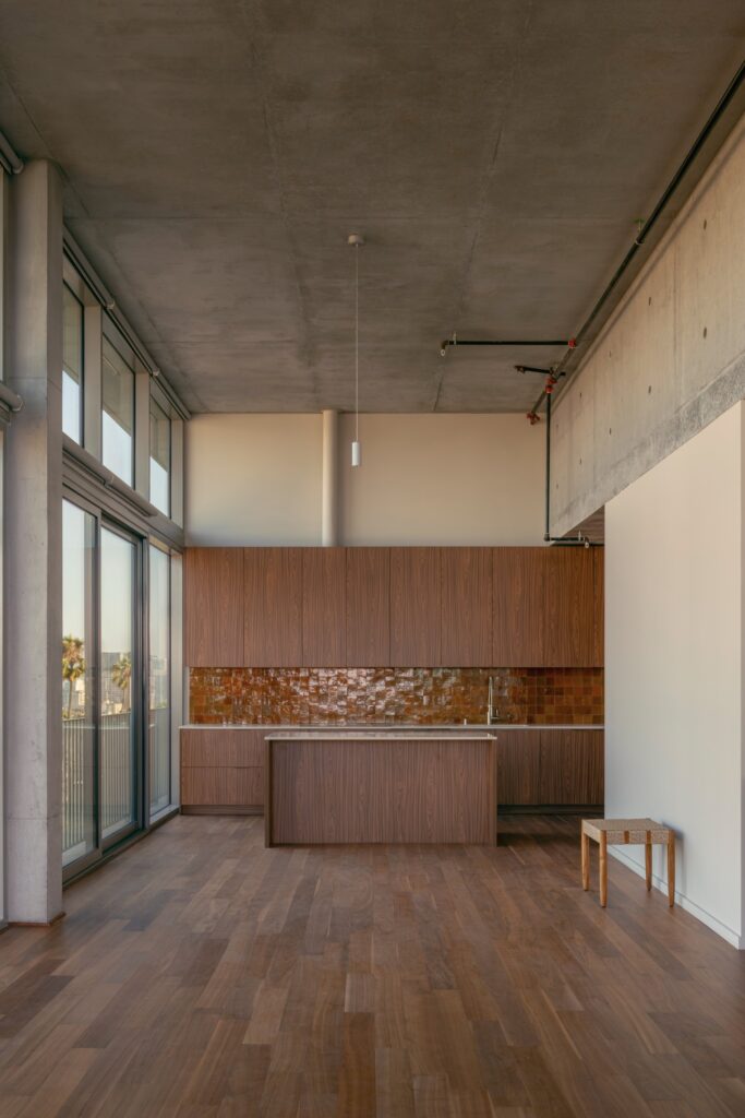Modern apartment interior in KAYA featuring a wooden kitchen island, glazed backsplash tiles, exposed concrete ceiling, and sliding glass doors.