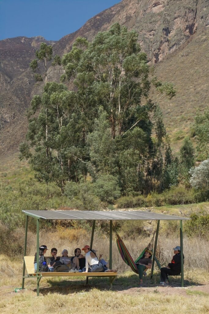 A group of people sitting under a lightweight open-air shaded metal structure with hammocks set against a steep mountain hillside.