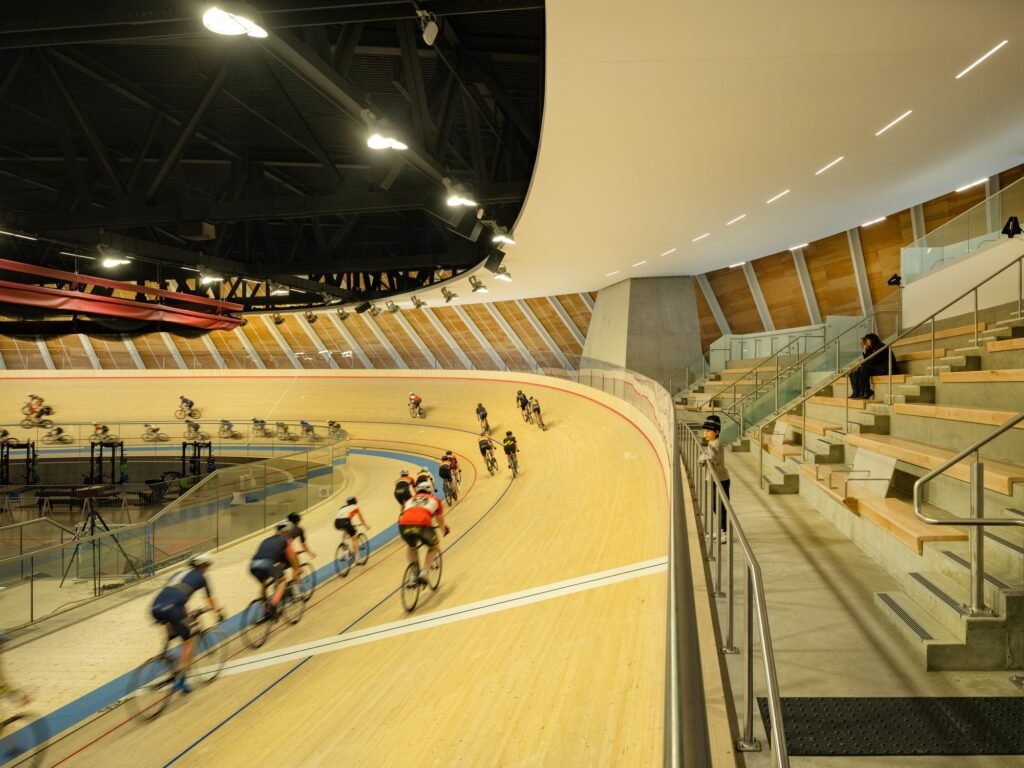 Cyclists navigating a steep banked turn on the wooden velodrome track with concrete spectator grandstand seating on the right.