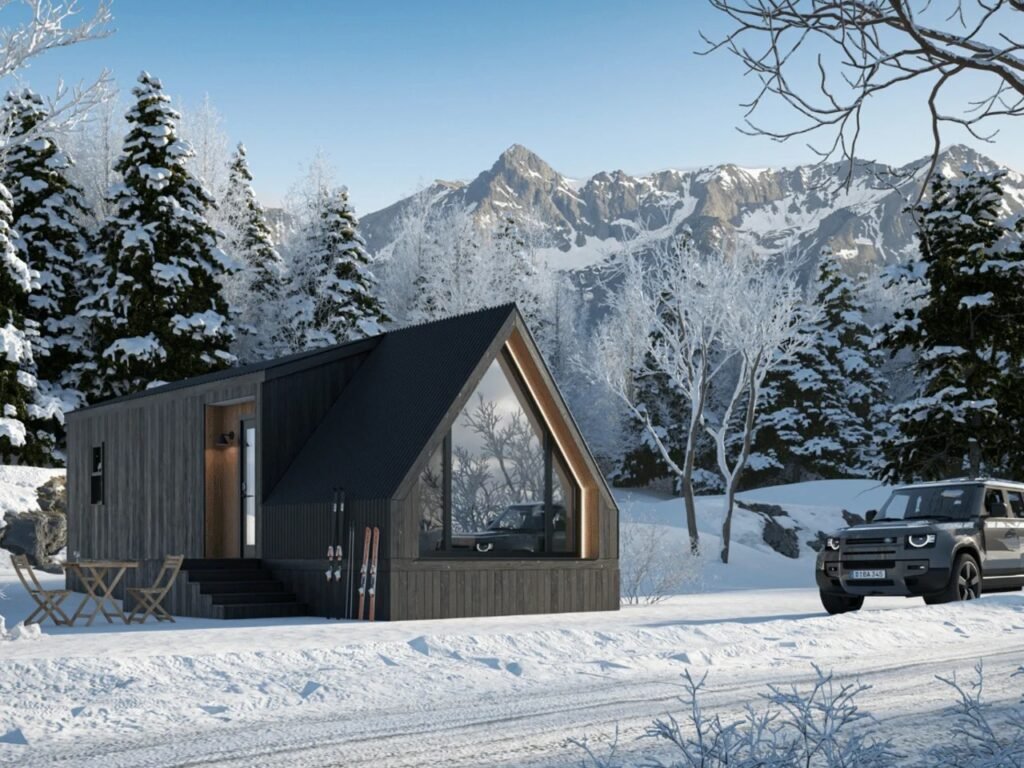 A-Frame Bunkhouse set in a snowy winter landscape with snow-covered pine trees, a mountain range background, and skis leaning against the matte black facade.