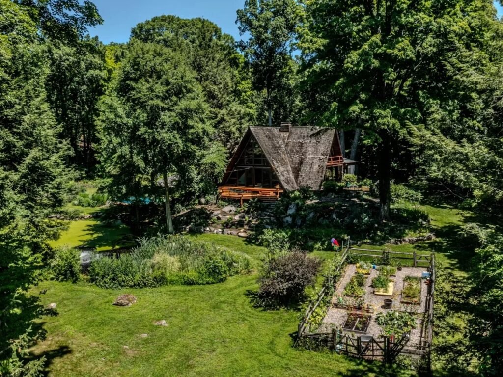 An aerial view of the Woods Home property, showing the main A-frame house surrounded by 6.74 acres of forest in Yorktown Heights. Below the house, a landscaped area includes a large circular garden plot enclosed by a low wooden fence, a small pond, and lawns. A swimming pool is visible near the house.