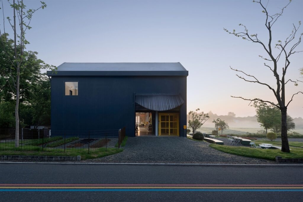 Ground-level exterior view at dusk of Spitfire Roastery's dark gable volume bordering a paved village road.