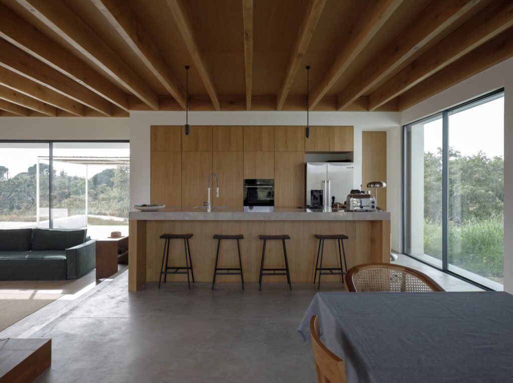 Open-plan kitchen and dining area inside Melides MG House featuring exposed wooden ceiling beams and large glazed windows.