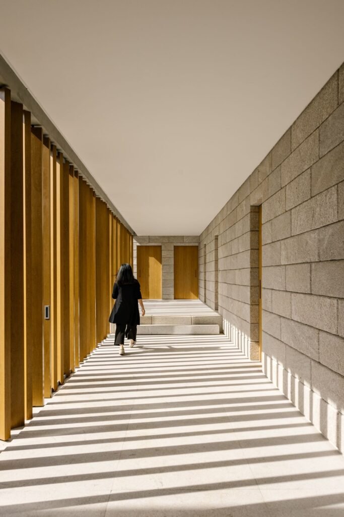 Person walking down a sunlit corridor bounded by a stone wall and vertical wooden louvers.