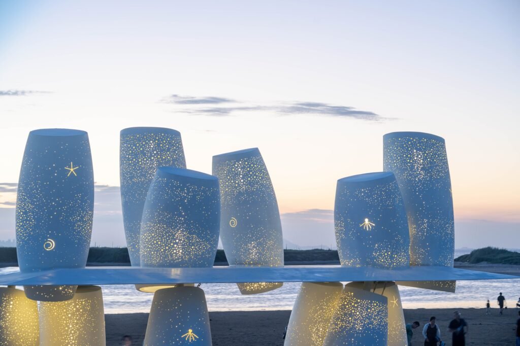 Detailed close-up of illuminated white steel cylinders at dusk, showing light filtering through dense patterns of circular and ocean-inspired cutouts.