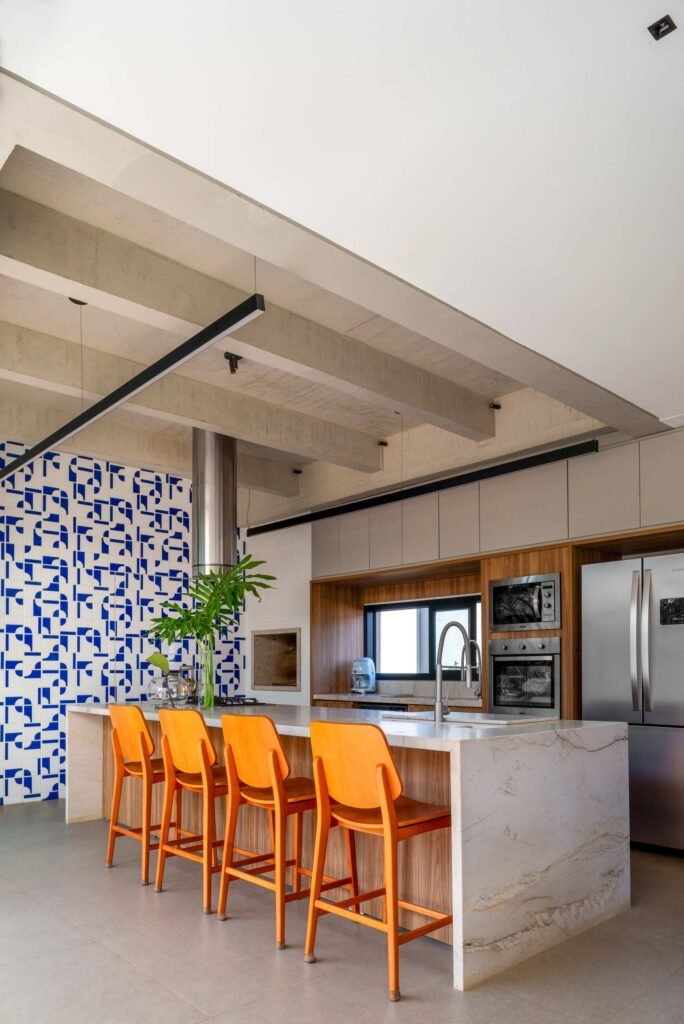 Open-plan kitchen in Casa Noor featuring exposed concrete ceiling beams, a marble island with orange stools, and a patterned tile wall.