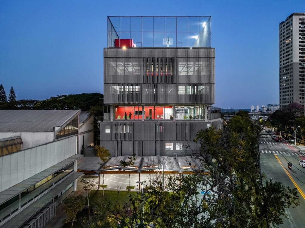 Exterior dusk view of illuminated High School building and adjacent industrial sheds with red interior accent volume visible.