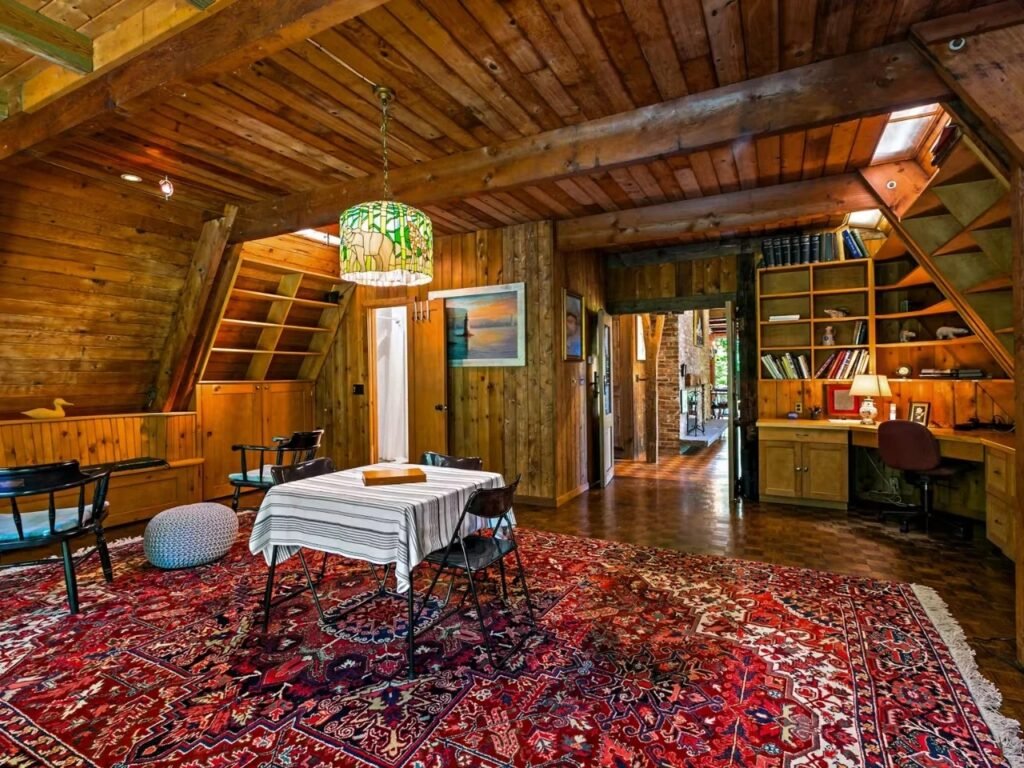 An interior view of the Woods Home living room, showcasing the vaulted, wood-paneled A-frame ceiling and exposed beams. A distinctive Tiffany-style pendant lamp with a nature pattern hangs above a table with a striped tablecloth and simple black chairs. An ornate red Persian-style rug covers the floor. Built-in wooden bookshelves and a desk area are visible on the right.