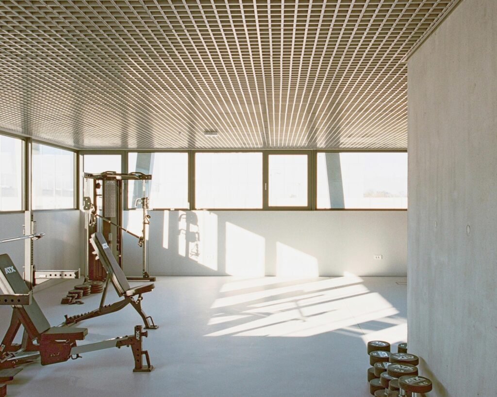 Interior view of a gym room with exercise equipment, dumbbells, a concrete core wall, expansive glass windows, and a metal mesh ceiling.