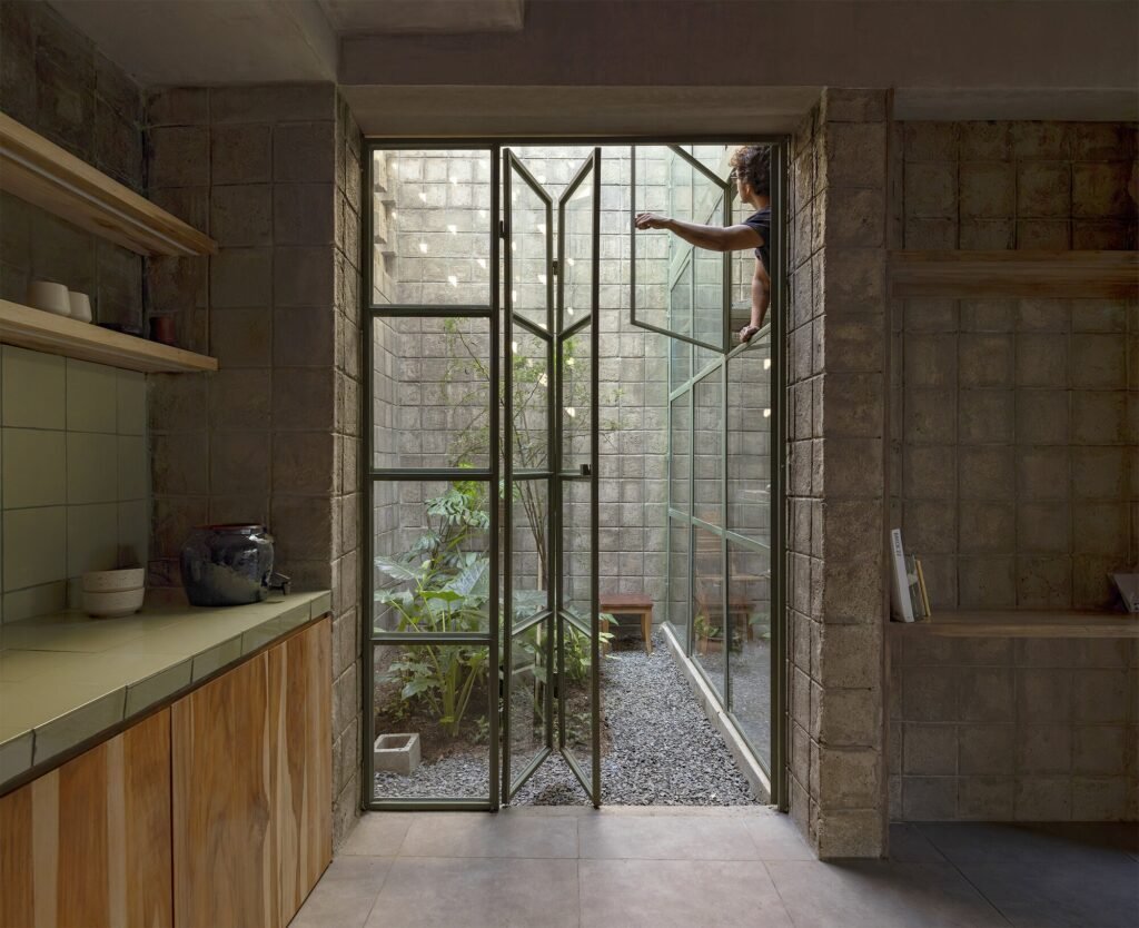 Interior kitchen looking out through folding steel-and-glass doors to an open-to-sky gravel patio with natural foliage, with a person seen on the upper level within the concrete block structure.