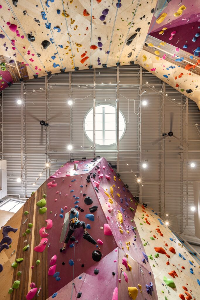 Upward perspective view of a climber ascending a purple climbing wall toward a central circular roof skylight.