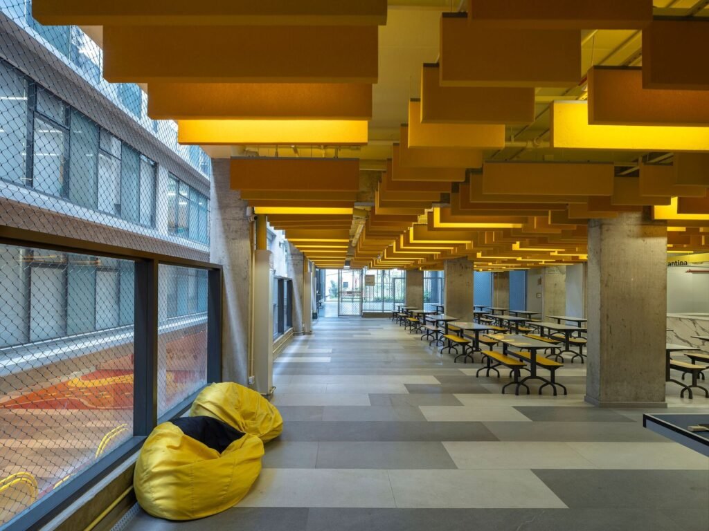 Bright student lounge and cafeteria area with yellow acoustic ceiling fins, bean bags, and dining tables next to a sports court.