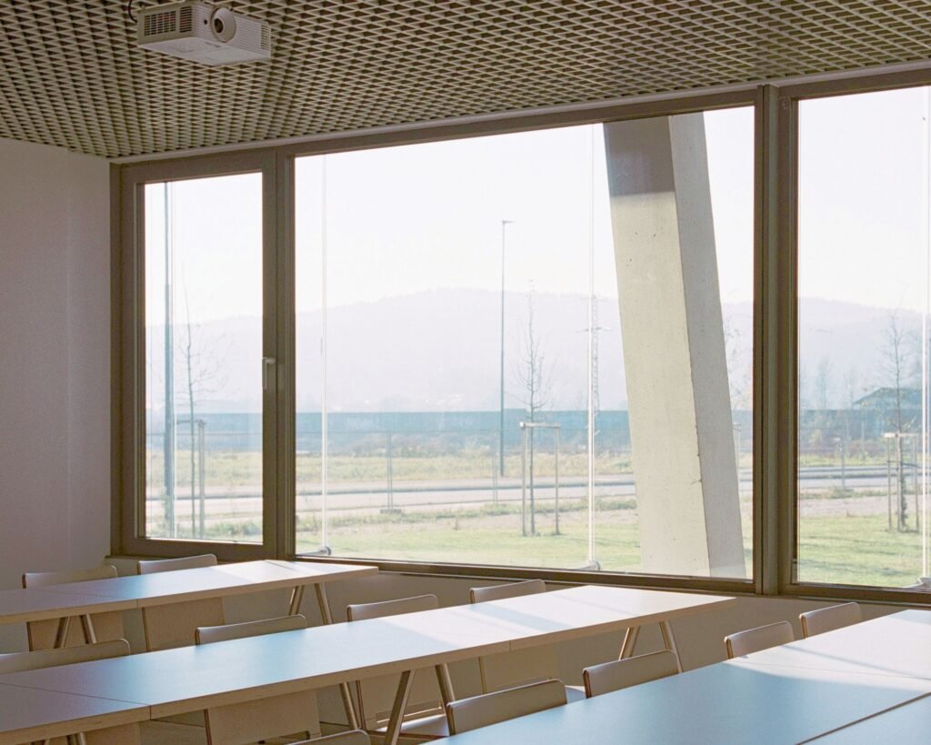 Interior of a bright educational classroom with wooden tables and chairs facing large full-height windows looking out onto the landscape and inclined column.